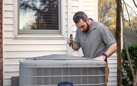A technician taking a reading at an outdoor condenser beside a house.