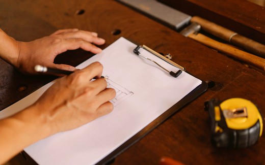 Hands writing on a clipboard on a workbench beside a tape measure.