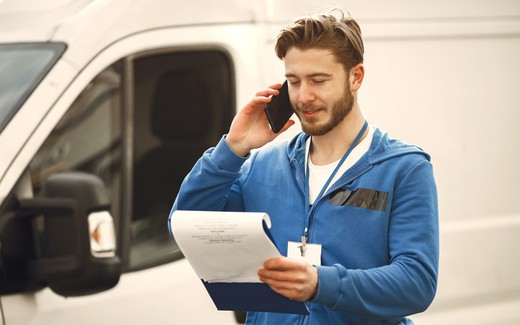 A man on a mobile phone beside a work van, clipboard in hand.
